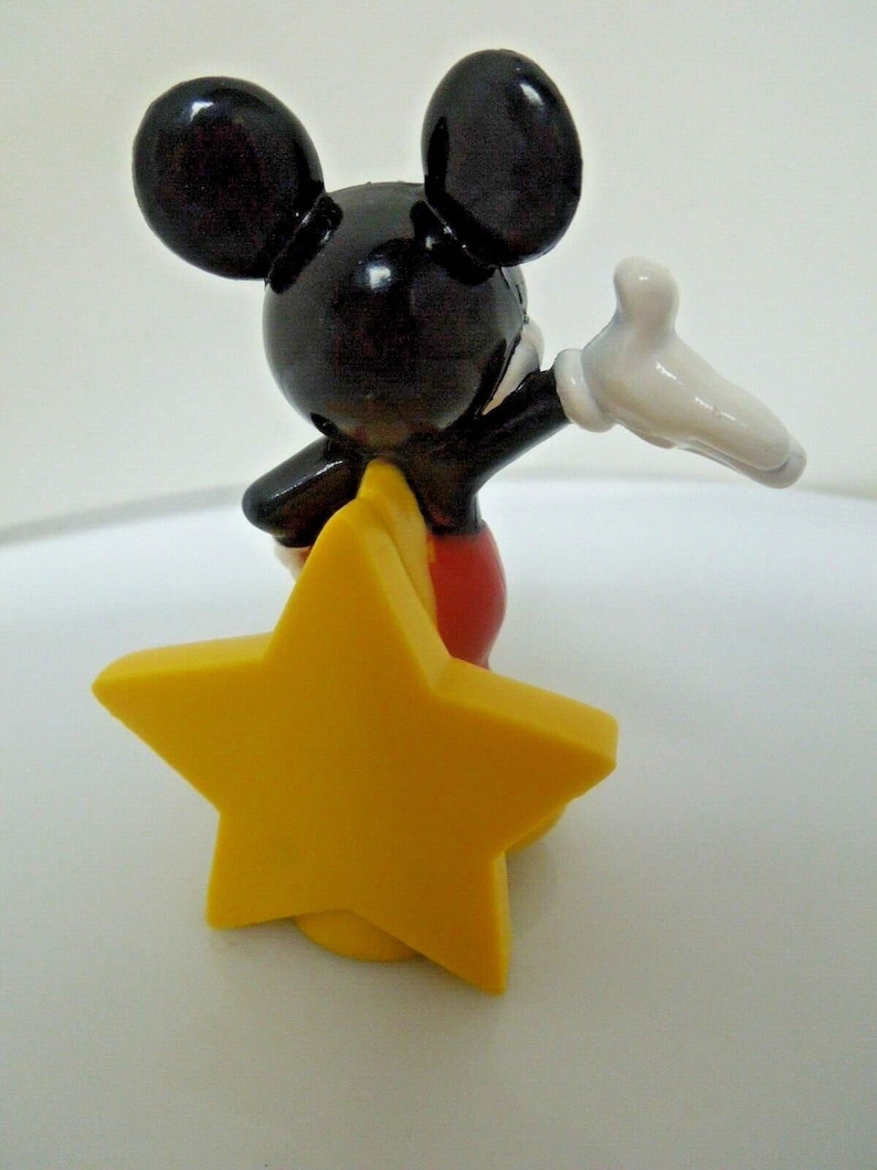 以下が含まれることがあります： 黄色い星の上に立っている黒と赤のプラスチック製のミッキーマウスのフィギュア。ミッキーマウスは右手で手を振っています。