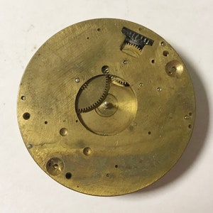 May include: A circular, brass-colored watch movement with intricate gears and small holes. The watch mechanism is a vintage piece, showing signs of age and wear. The gears are visible, and the overall design is detailed and complex.