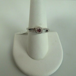 May include: A silver ring with a red gemstone surrounded by smaller white gemstones. The ring is on a white ring display stand.