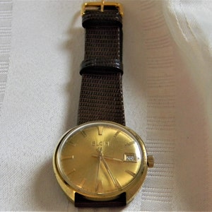May include: A gold-toned Elgin wristwatch with a brown leather band. The watch face has a date window and the text "Elgin" printed on it.