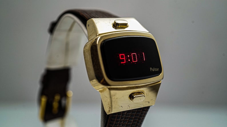 May include: A gold-toned Pulsar digital watch with a black face and red LED display showing the time 9:01. The watch has a brown leather strap.