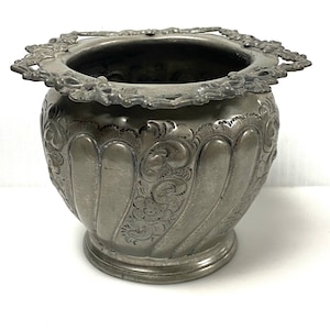 May include: A metallic silver-toned bowl with a wide, ornate, scalloped rim featuring intricate floral engravings. The body of the bowl has vertical fluted panels alternating with detailed etched scrollwork and flower patterns, resting on a circular base.