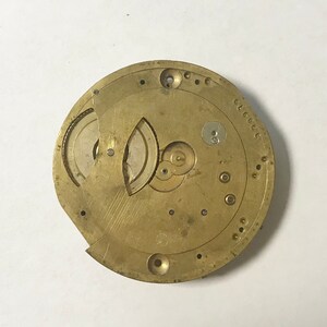 May include: A gold-colored watch movement with intricate gears and a circular design. The movement is disassembled and shows the inner workings of the watch.