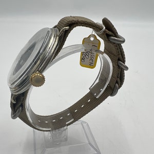 May include: A silver wristwatch with a black face and a brown canvas strap. The watch has a gold crown and is on a clear display stand. A yellow tag with the text "WISSEN 1950" is attached to the strap.