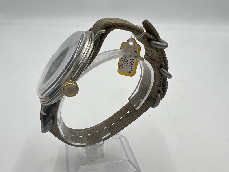 May include: A silver wristwatch with a black face and a brown canvas strap. The watch has a gold crown and is on a clear display stand. A yellow tag with the text "WISSEN 1950" is attached to the strap.