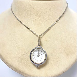 May include: A silver chain necklace with a silver pocket watch pendant. The watch face is white with black Roman numeral hour markers and black hands.