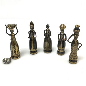 May include: A collection of five antique brass figurines depicting stylized figures in various poses. The figurines have a dark patina with gold accents, showcasing intricate details. One figure carries a basket on its head, while others hold objects or have their arms crossed.