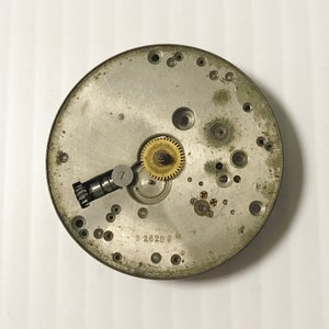 May include: Close-up of a silver watch movement with a gold gear. The movement has a number of small holes and a number "3 2826 9" printed on it.