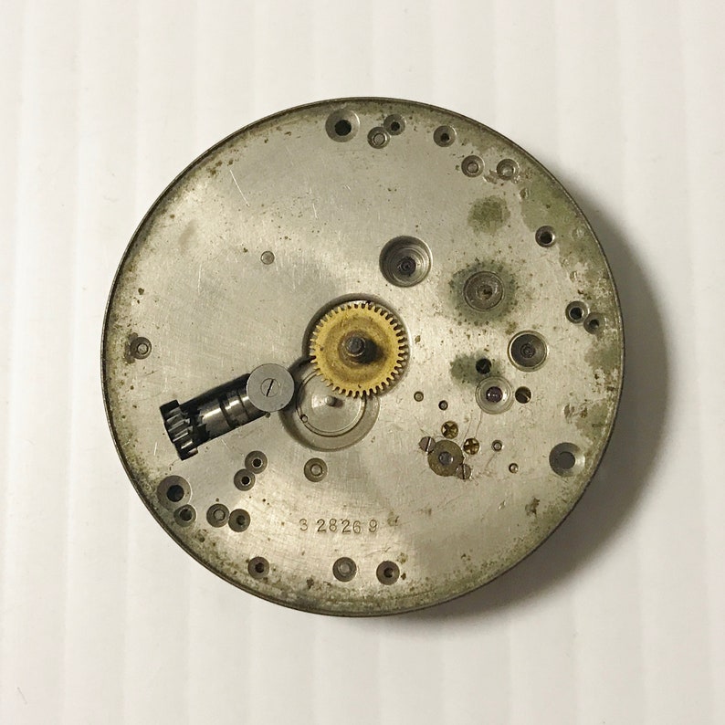 May include: Close-up of a silver watch movement with a gold gear. The movement has a number of small holes and a number "3 2826 9" printed on it.