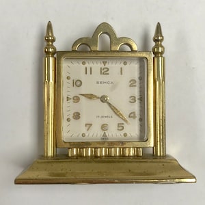 May include: A gold-toned, vintage, tabletop clock with a white face and black hands. The clock face reads "SEMCA 17 JEWELS SWISS".
