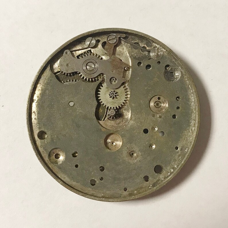May include: Close-up view of the inner workings of a silver-toned watch mechanism. The intricate gears and levers are visible, showcasing the complex design of the watch.