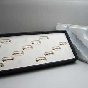 May include: A white ring display tray with black edging and multiple ring-shaped indentations. The tray is sitting on a white surface.