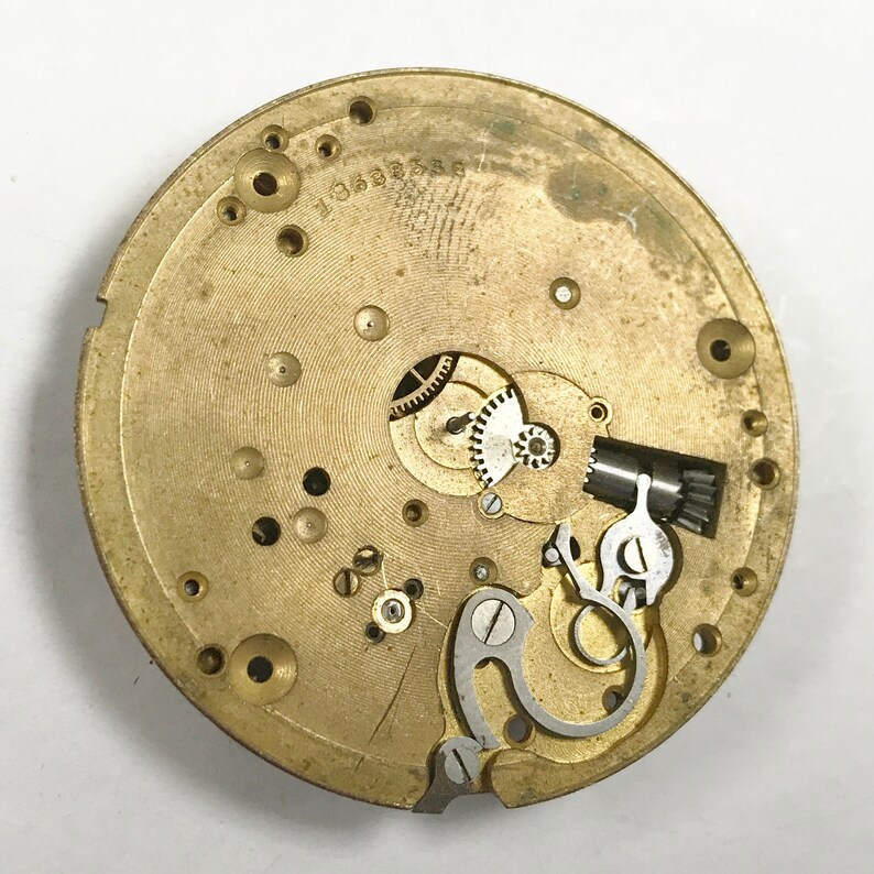 K&ouml;nnte beinhalten: Nahaufnahme eines goldfarbenen Uhrwerks mit komplizierten Zahnr&auml;dern und einem Aufzugsmechanismus. Das Uhrwerk ist durch den Geh&auml;useboden der Uhr sichtbar.