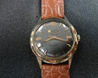 May include: A gold-tone wristwatch with a black face and a brown leather band. The watch face has gold-tone hands and markers, and the text "Sea-shark Automatic" is printed on the face.