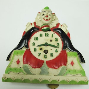 May include: A vintage clown clock with a red, white, and green color scheme. The clock face is white with black numbers and hands. The clown is holding a red and green ball in its hand. The clock is hanging from a hook.