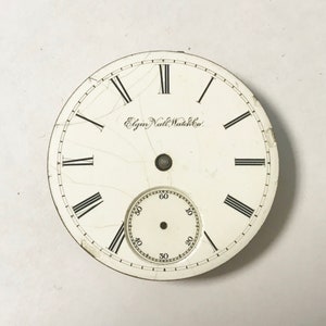 May include: White watch face with black Roman numeral hour markers and black minute markers. The watch face has a small, white sub-dial with black numbers. The text "Elgin Natl. Watch Co." is printed on the watch face.
