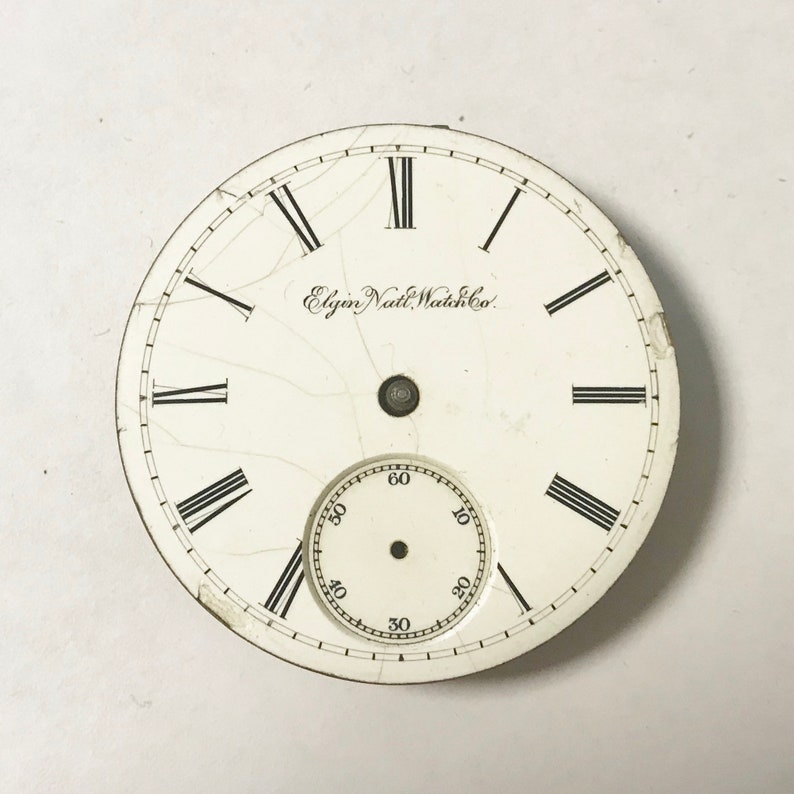 May include: White watch face with black Roman numeral hour markers and black minute markers. The watch face has a small, white sub-dial with black numbers. The text "Elgin Natl. Watch Co." is printed on the watch face.