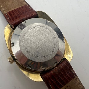 Puede incluir: Parte posterior de un reloj de pulsera dorado con una correa de cuero marr&oacute;n. La esfera del reloj es de acero inoxidable y tiene el texto "STAINLESS STEEL BACK  WATER RESISTANT  AUTOMATIC  ROCALOC  17 JEWELS  SWISS MADE" grabado.