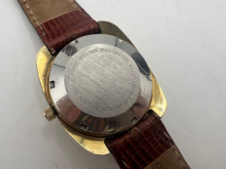 Puede incluir: Parte posterior de un reloj de pulsera dorado con una correa de cuero marr&oacute;n. La esfera del reloj es de acero inoxidable y tiene el texto "STAINLESS STEEL BACK  WATER RESISTANT  AUTOMATIC  ROCALOC  17 JEWELS  SWISS MADE" grabado.