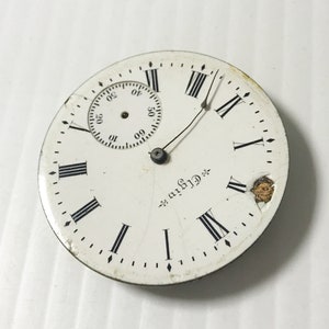 May include: A white pocket watch face with black Roman numeral hour markers and a smaller, circular sub-dial. The watch face is cracked and has a small piece of metal missing. The text "Griffin" is printed on the watch face.