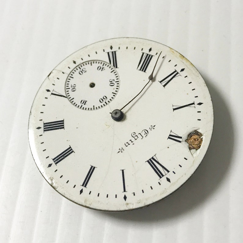May include: A white pocket watch face with black Roman numeral hour markers and a smaller, circular sub-dial. The watch face is cracked and has a small piece of metal missing. The text "Griffin" is printed on the watch face.