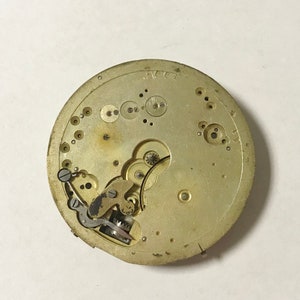 K&ouml;nnte beinhalten: Nahaufnahme eines goldfarbenen Uhrwerks mit komplizierten Zahnr&auml;dern und einer silberfarbenen Aufzugsfeder. Das Uhrwerk ist vor einem wei&szlig;en Hintergrund platziert.