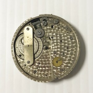May include: Close-up of a silver watch movement with intricate gears and a textured surface. The number 15489 is visible on the bottom of the movement.
