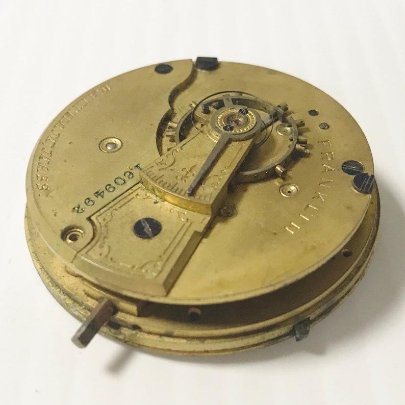 May include: A gold-toned pocket watch movement with intricate gears and a winding key. The watch face is marked "FRANKLIN" and has the number "2646091".