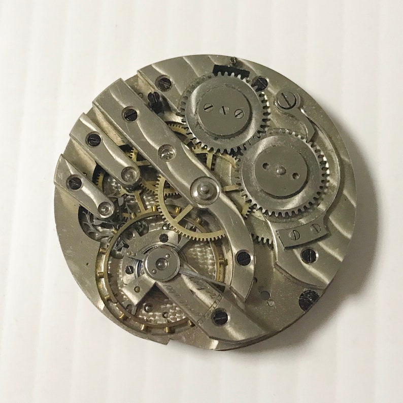 May include: Close-up of a silver pocket watch movement with intricate gears and a winding stem. The watch face is not visible.