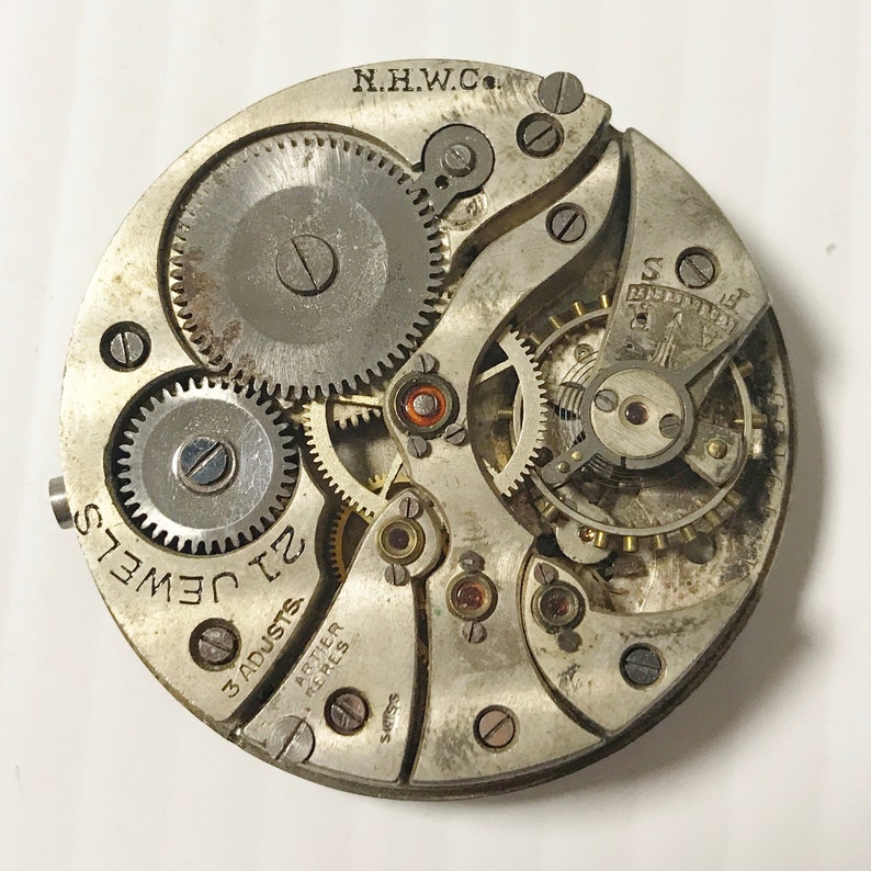 May include: Close-up of a silver watch movement with the text "N.H.W.C." and "21 Jewels" visible. The movement has a number of gears and other components, including a lever with the text "3 Adjusts" and "Arter Peres" visible.