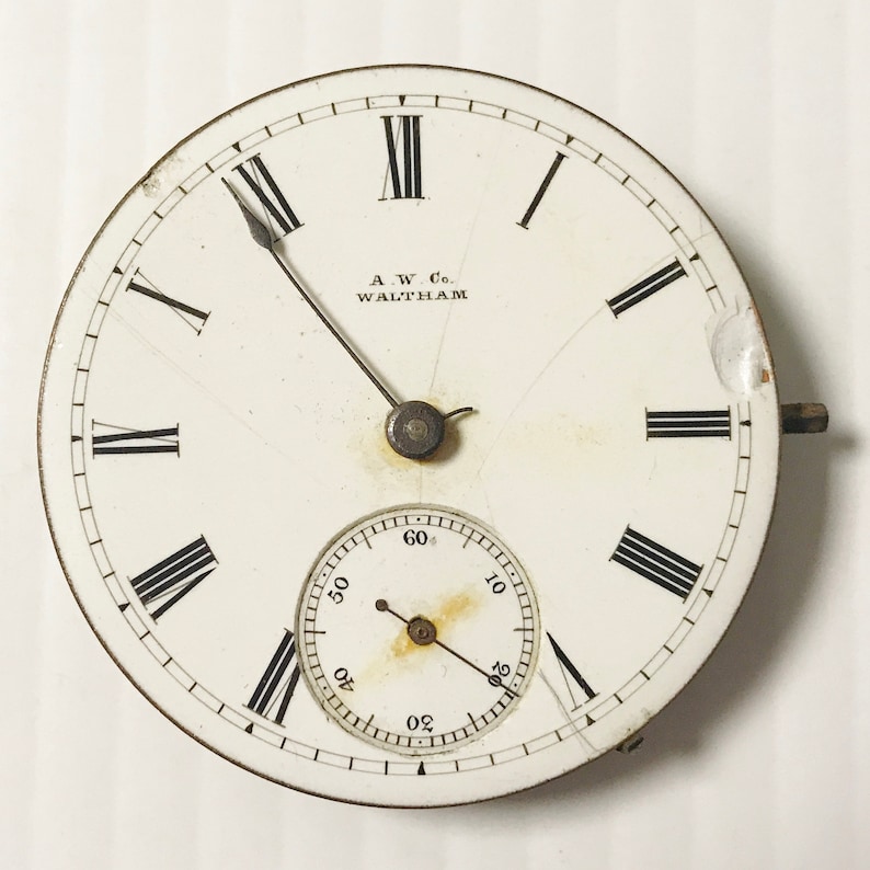 May include: Antique pocket watch face with a white background, black Roman numerals, and a smaller seconds dial. The watch face has the text "A.W. Co. WALTHAM" and shows signs of age and wear.
