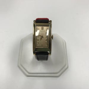 May include: A gold-toned Waltham wristwatch with a rectangular face and black leather band. The watch face has white Roman numeral hour markers and black hands.