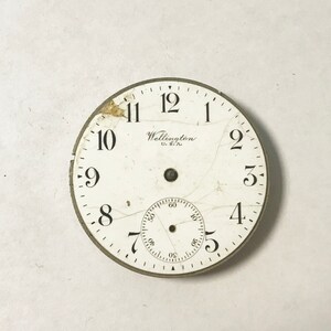 以下が含まれることがあります： 黒色の数字と針が付いた白い時計の文字盤。文字盤はひび割れており、中央に小さな円形の穴があります。文字盤には「Wellington U.S.A.」という文字が印刷されています。