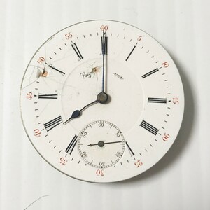 Puede incluir: Un reloj de bolsillo blanco con marcadores de hora romanos negros y agujas negras. La esfera del reloj tiene un peque&ntilde;o subdial circular con agujas negras y marcas de n&uacute;meros ar&aacute;bigos. La esfera del reloj est&aacute; agrietada y tiene las palabras "Eng" y "ver" impresas.