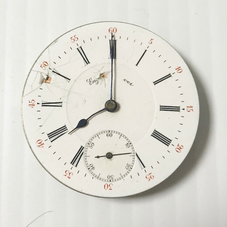 Puede incluir: Un reloj de bolsillo blanco con marcadores de hora romanos negros y agujas negras. La esfera del reloj tiene un peque&ntilde;o subdial circular con agujas negras y marcas de n&uacute;meros ar&aacute;bigos. La esfera del reloj est&aacute; agrietada y tiene las palabras "Eng" y "ver" impresas.