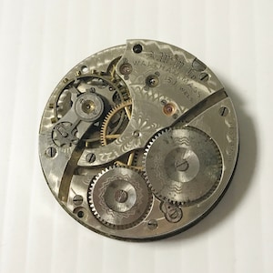 May include: Close-up of a silver pocket watch movement with intricate gears and a decorative floral pattern. The watch is labeled "A.W. Co. Waltham, Mass. 15J. Jewels."