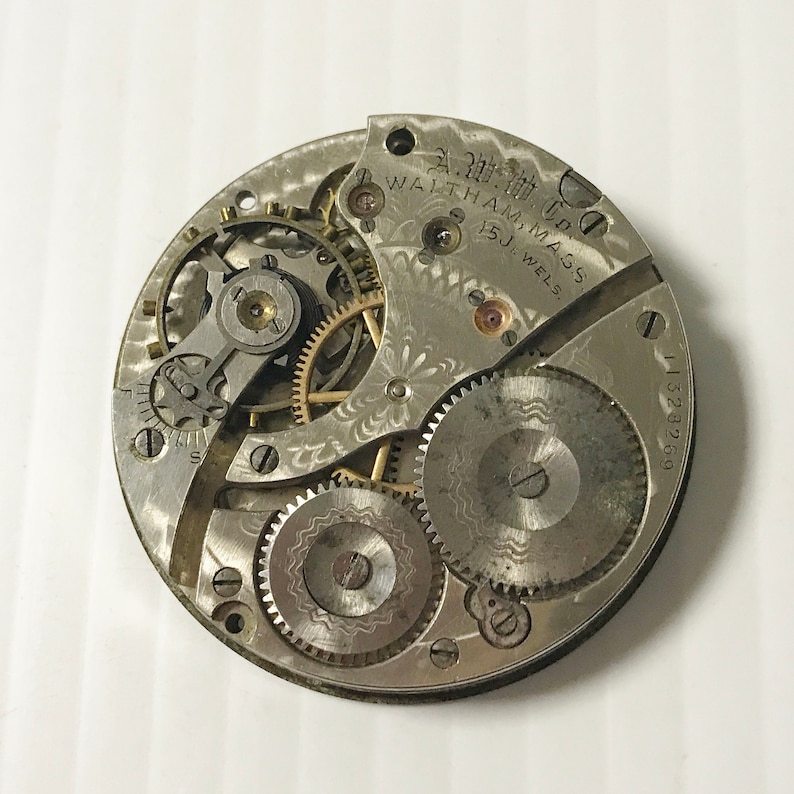 May include: Close-up of a silver pocket watch movement with intricate gears and a decorative floral pattern. The watch is labeled "A.W. Co. Waltham, Mass. 15J. Jewels."
