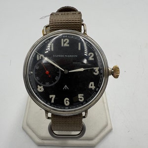 May include: A black-faced wristwatch with white numerals and hands. The watch has a silver case and a brown canvas strap. The watch face reads "ULYSSE NARDIN".