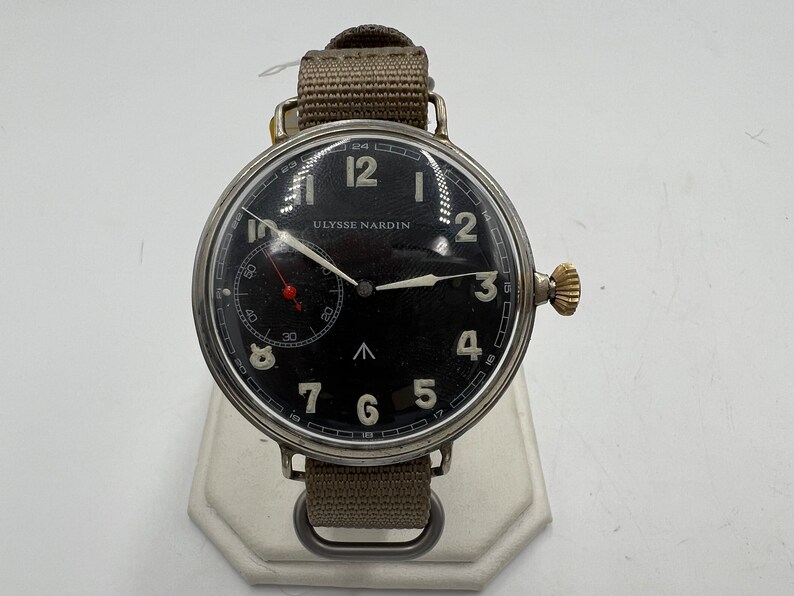 May include: A black-faced wristwatch with white numerals and hands. The watch has a silver case and a brown canvas strap. The watch face reads "ULYSSE NARDIN".