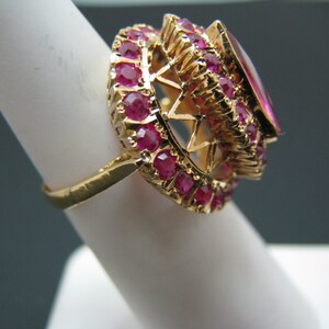 May include: A gold ring with a large, oval-shaped ruby surrounded by a halo of smaller rubies.