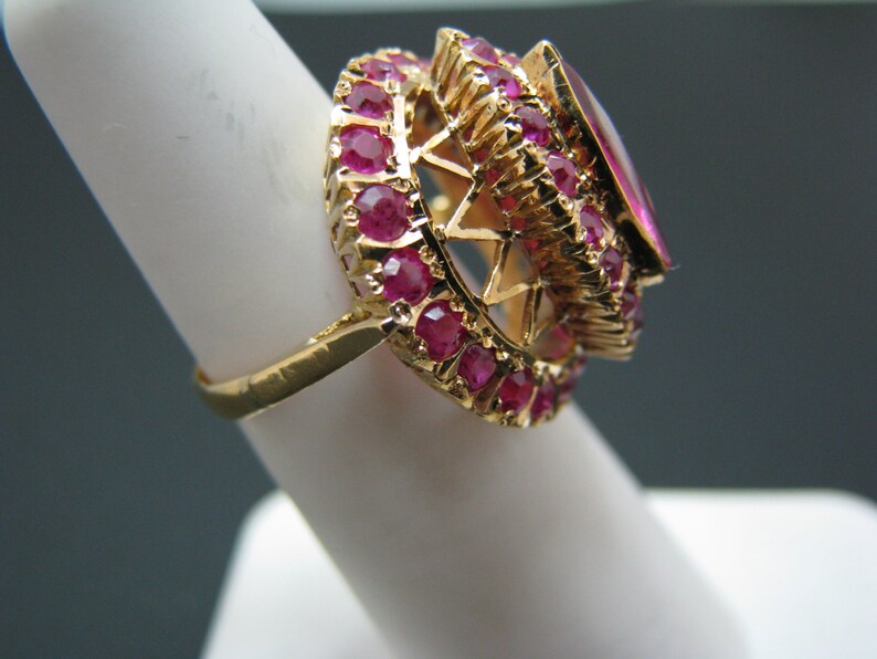 May include: A gold ring with a large, oval-shaped ruby surrounded by a halo of smaller rubies.