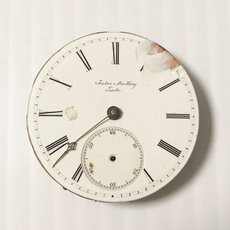 May include: White pocket watch face with black Roman numeral hour markers and black markings for the seconds. The watch face has the text "Jules Mahey ecole" in black script.