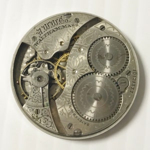 May include: Close-up of a vintage watch movement. The silver-toned mechanism features intricate gears, springs, and decorative engravings. The text "A.W.W.Co. WALTHAM, MASS." is visible. The watch part has a serial number "10121377".