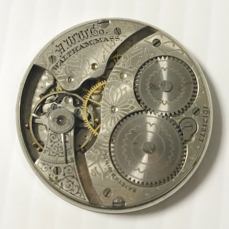 May include: Close-up of a vintage watch movement. The silver-toned mechanism features intricate gears, springs, and decorative engravings. The text "A.W.W.Co. WALTHAM, MASS." is visible. The watch part has a serial number "10121377".