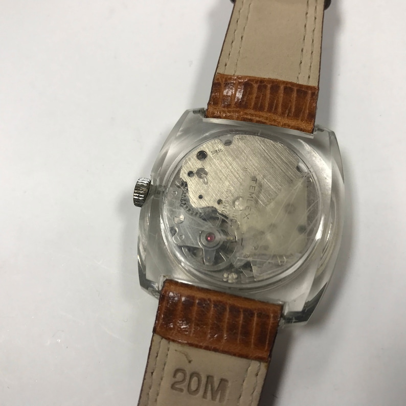 Peut inclure: Une montre carr&eacute;e transparente avec un bracelet en cuir marron. Le cadran de la montre est visible &agrave; travers le bo&icirc;tier transparent, montrant le fonctionnement interne de la montre. Le bracelet porte l'inscription "20M".