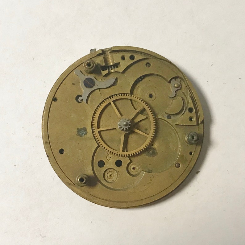 May include: A close-up of a gold-toned watch movement with intricate gears and a large central gear. The movement is disassembled and laid out on a white surface.