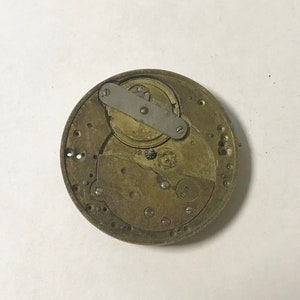 May include: Close-up of a gold-toned watch movement with a silver-toned lever and gears. The movement is circular and has a number of holes around the edge.