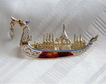 May include: A silver and enamel brooch in the shape of a traditional Thai longtail boat. The boat is decorated with intricate details and features a red, white, and blue color scheme.