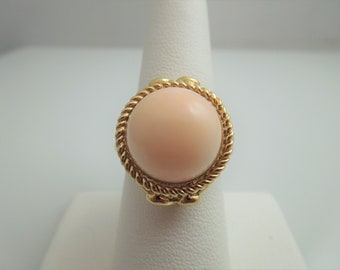 May include: A gold ring featuring a large, round, light pink gemstone. The gemstone is surrounded by a twisted rope-like gold band. The ring is displayed on a white ring holder.