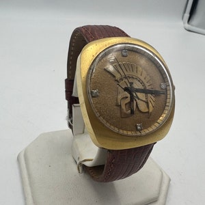 Puede incluir: Un reloj de pulsera dorado con una correa de cuero marr&oacute;n. La esfera del reloj presenta una esfera romana dorada con agujas doradas de hora y minuto. La esfera del reloj tambi&eacute;n presenta una imagen dorada de un casco de guerrero.